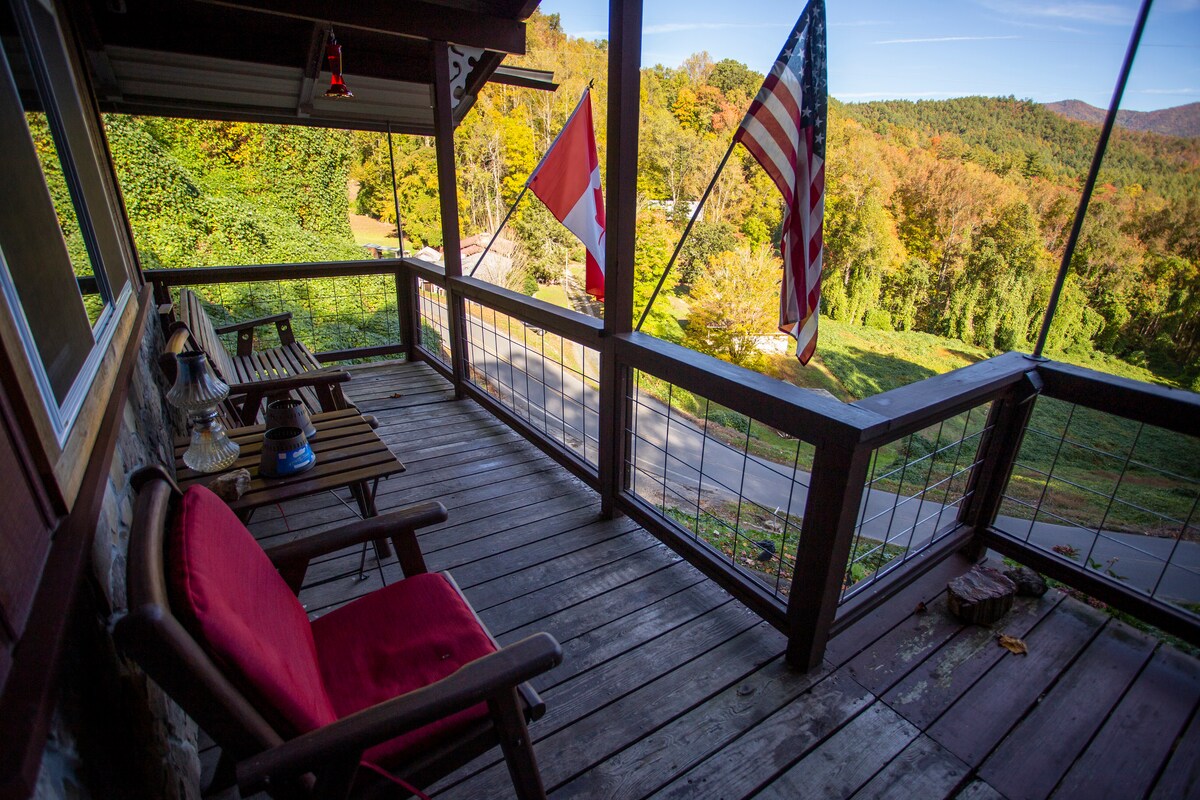 A wraparound deck features wooden flooring and is furnished with a red cushioned chair and a small table. Two flags, one Canadian and one American, are visibly displayed. The surrounding area showcases vibrant trees, indicating a colorful foliage season, with mountains in the distance.