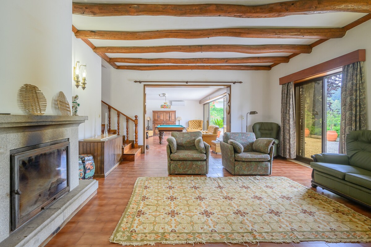A spacious living area features wooden beams overhead and a large patterned rug covering the floor. Two armchairs and a reclining chair are arranged thoughtfully, while natural light enters through sliding glass doors leading to the garden. A fireplace creates a cozy focal point.