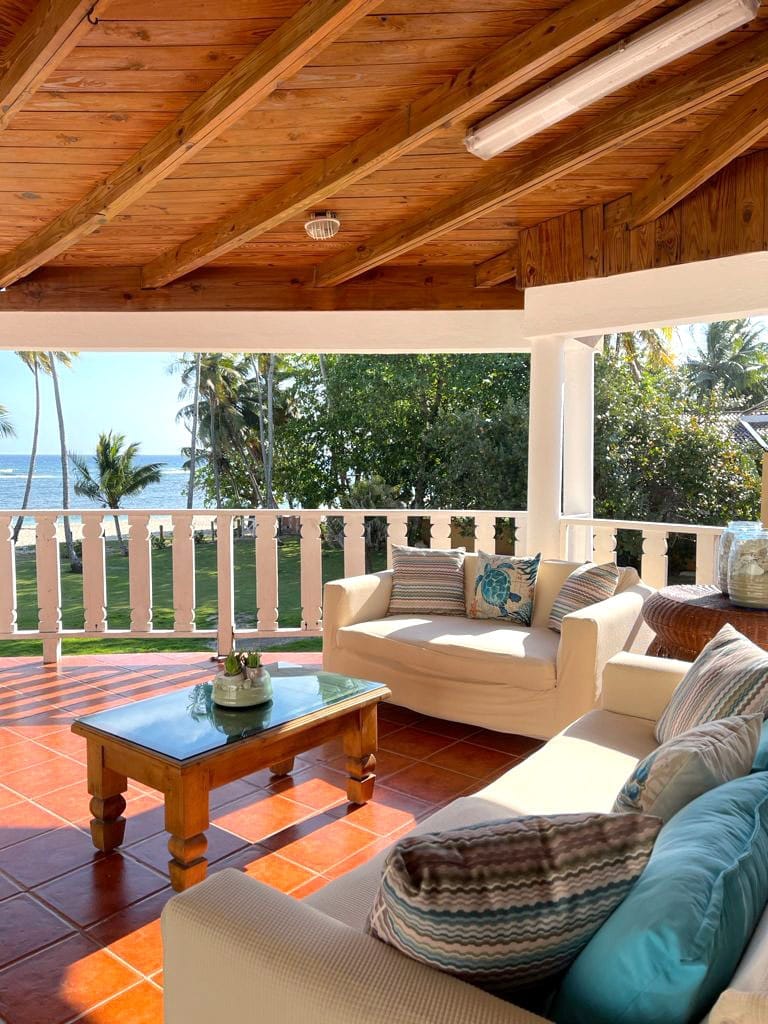 A comfortable seating area features a light-colored sofa adorned with decorative pillows, positioned around a wooden coffee table. The space is enhanced by a slatted wooden ceiling and offers views of the ocean and surrounding greenery through the open balcony railing.