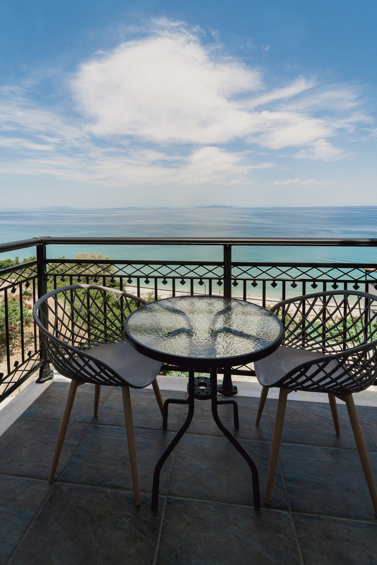 An outdoor balcony is depicted, featuring two modern chairs surrounding a round glass-topped table. The railing offers a view of the sea under a clear blue sky, contributing to a serene coastal atmosphere.