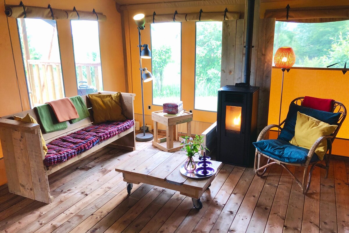 A spacious seating area is furnished with a wooden sofa adorned with colorful blankets and plush cushions. A stylish lamp stands beside a small table, while a cozy sitting chair complements the decor. A wood stove adds warmth, enhancing the inviting ambiance of the tent.