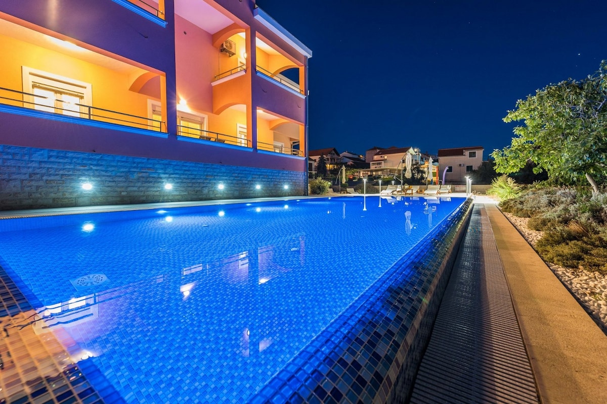 The illuminated infinity pool reflects a deep blue hue against the evening sky. Lights along the pool's edge provide a serene ambiance, while the surrounding area features landscaped gardens and a pathway leading up to the vibrant building.