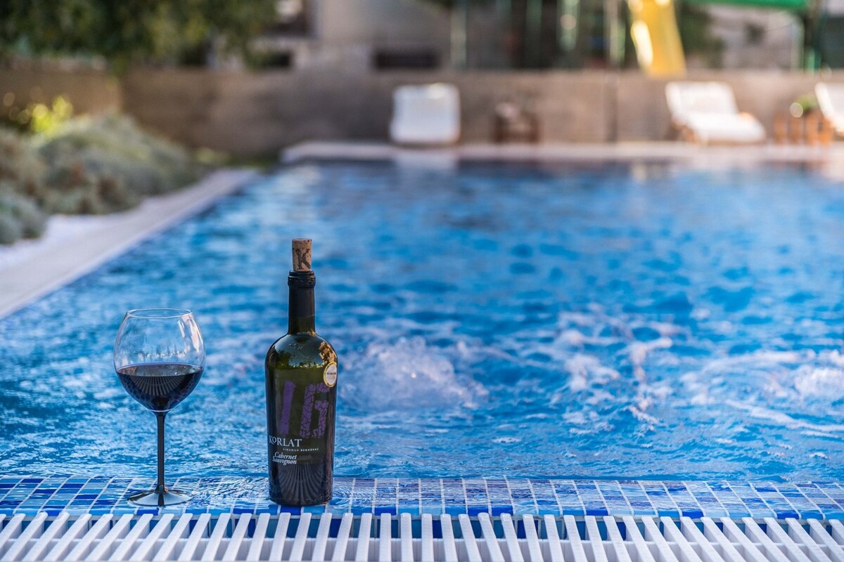 A bottle of red wine and a glass are positioned on the edge of a vibrant blue infinity pool. The pool's surface reflects sunlight while hydro-massage jets create gentle ripples, enhancing the relaxing atmosphere.