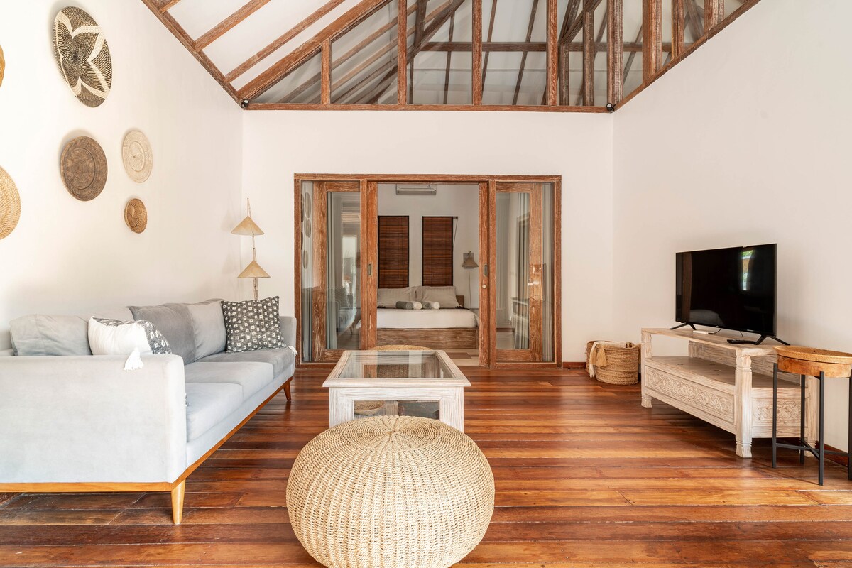 The living room features a spacious layout with wooden flooring and a high ceiling supported by exposed beams. A light grey sofa is positioned near a glass coffee table, while a flat-screen TV is mounted on a wooden console. Sliding glass doors lead to the bedroom area.