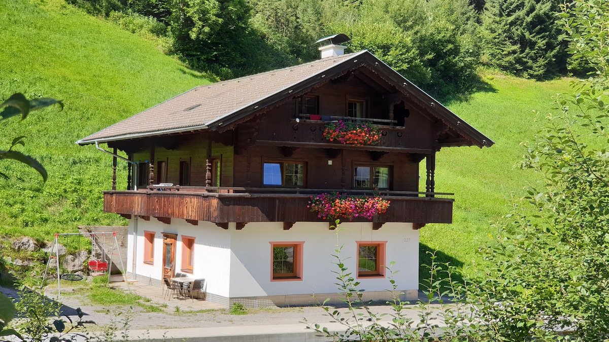 A charming two-story chalet is situated against a backdrop of lush green hills. Wooden balconies adorned with colorful flower boxes enhance the exterior. Large windows reflect natural light, while a porch area with seating invites relaxation. The building is surrounded by greenery, creating a serene atmosphere.