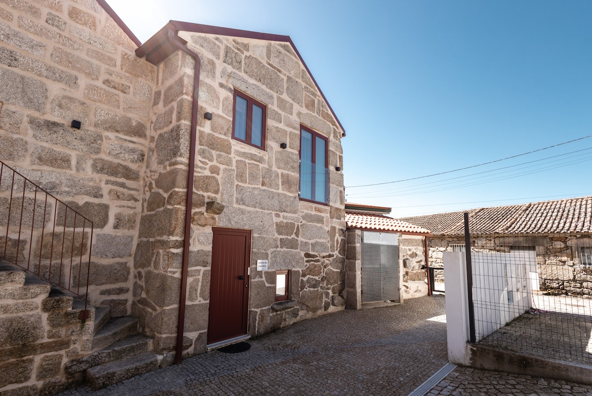 A two-story stone building features large windows and a dark red door. The textured stone facade contrasts with the clear blue sky. An outdoor area is visible, including a gravel path leading to a fenced section of the property.