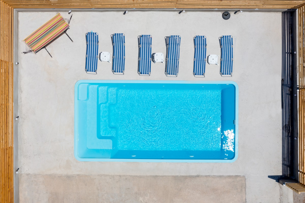 An aerial view captures a private swimming pool bordered by sun loungers featuring blue and white striped fabric. A colorful striped parasol is positioned beside the pool, providing shade. The surface surrounding the pool is smooth and light, emphasizing the vibrant blue water.