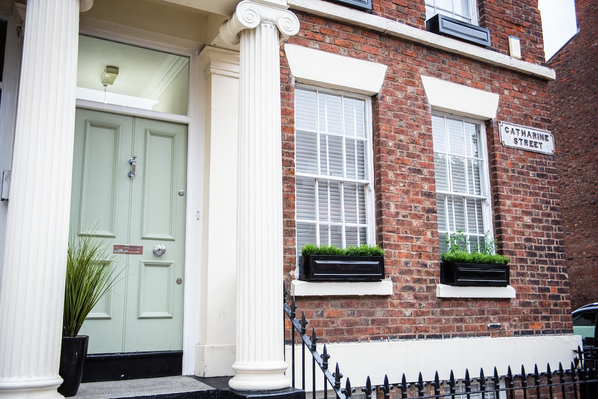 A historic entrance to the townhouse features tall white columns framing a wooden door painted in a soft green shade. Flower boxes with greenery sit below the windows of the brick facade, and a street sign indicates Catharine Street.