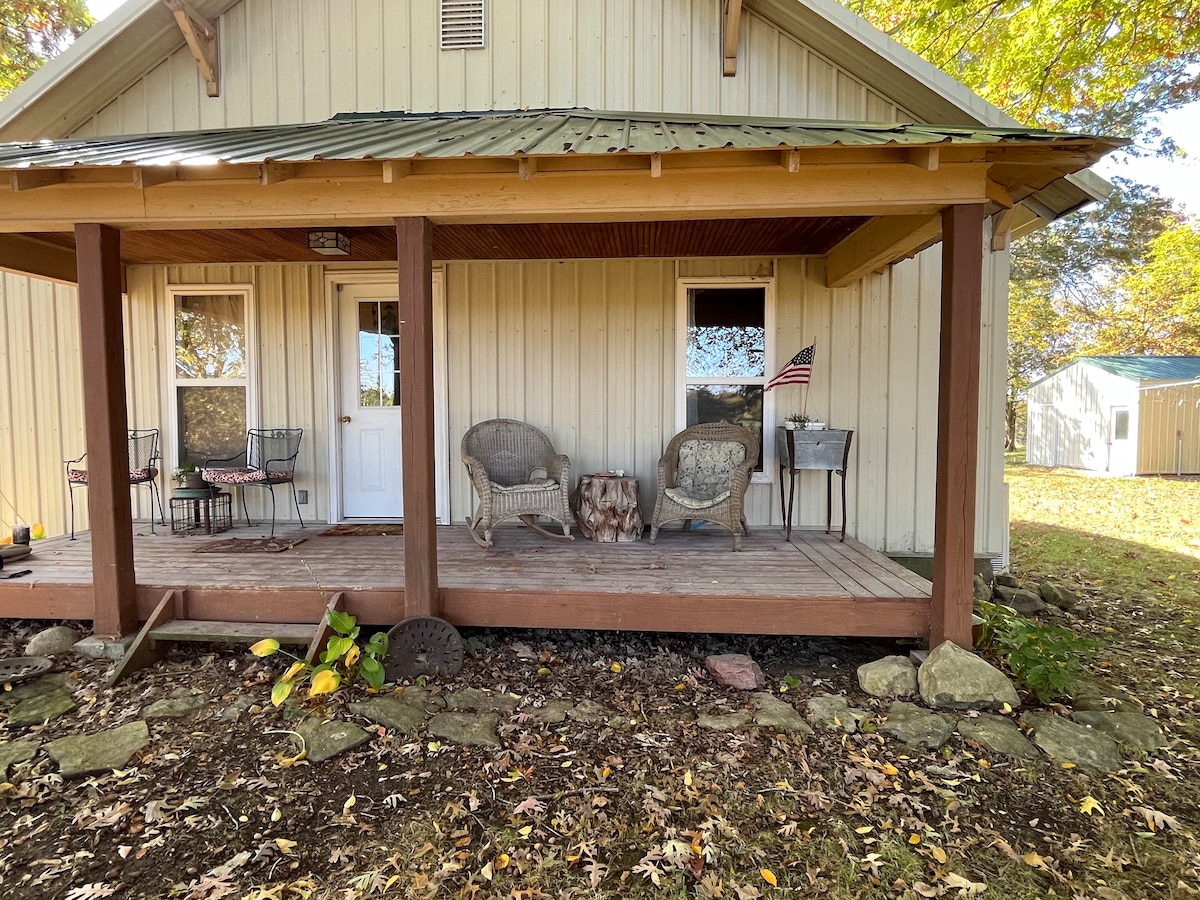 A shaded front porch features two vintage chairs and a small table. An American flag decorates one chair. The wooden decking is flanked by natural stone, with autumn leaves scattered on the ground. Two windows provide light to the entrance.