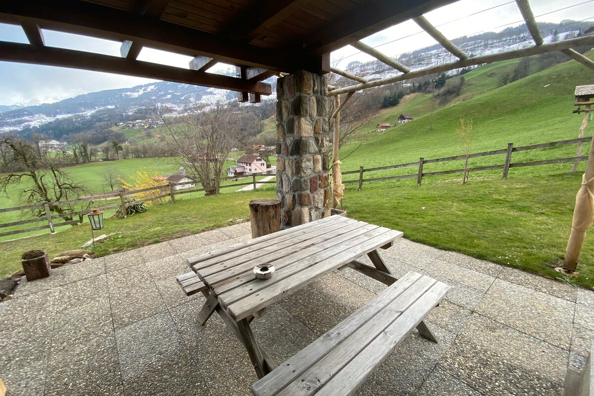 A wooden picnic table with benches is placed under a covered terrace, overlooking a large green meadow. A stone pillar supports the roof, and distant hills are visible, enhancing the serene outdoor setting.