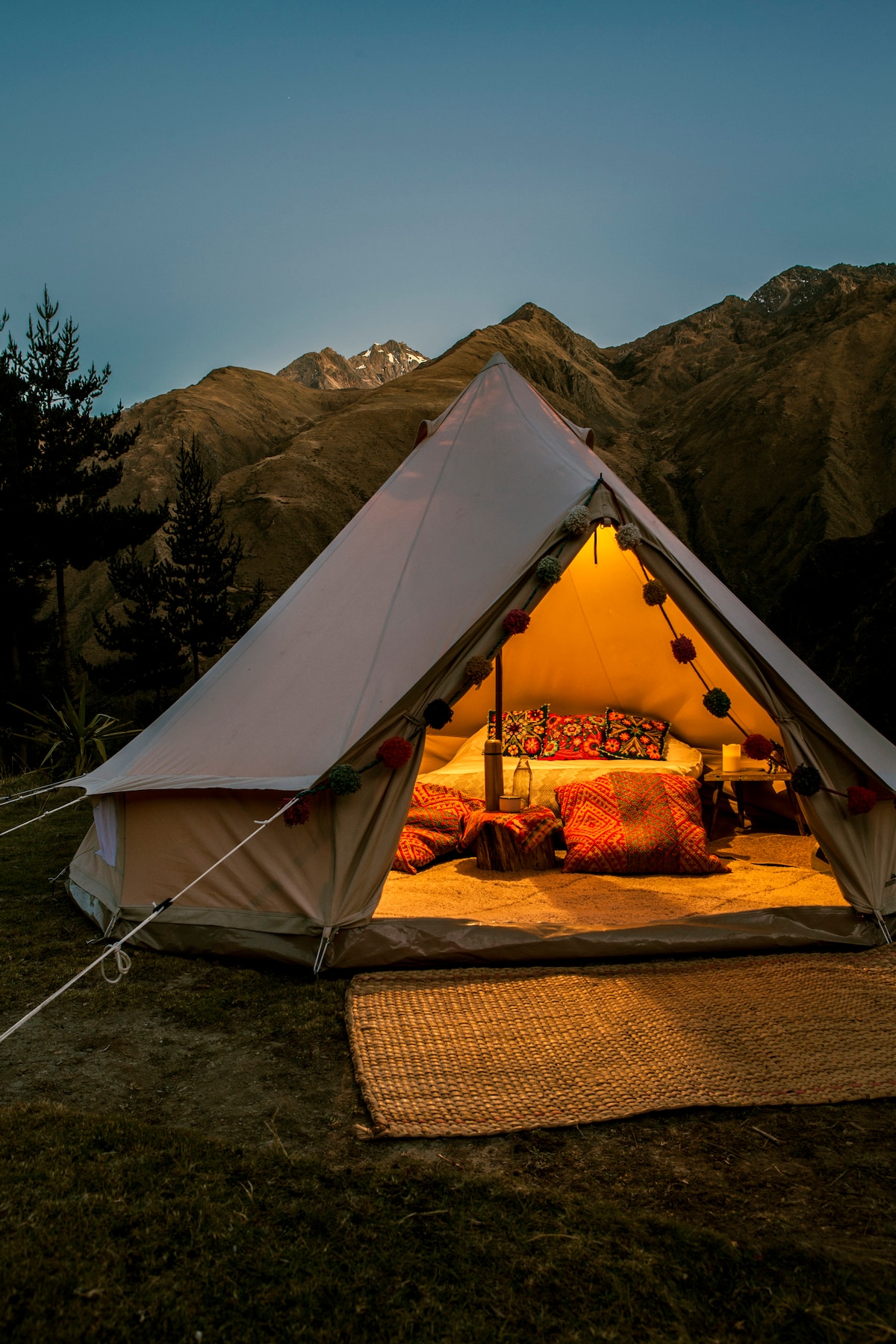 A spacious glamping tent is set against a mountainous backdrop, illuminated by soft, warm lighting. Inside, vibrant cushions are arranged on a plush rug, enhancing the inviting atmosphere. The tent's exterior is adorned with decorative elements, creating a cozy retreat in nature.