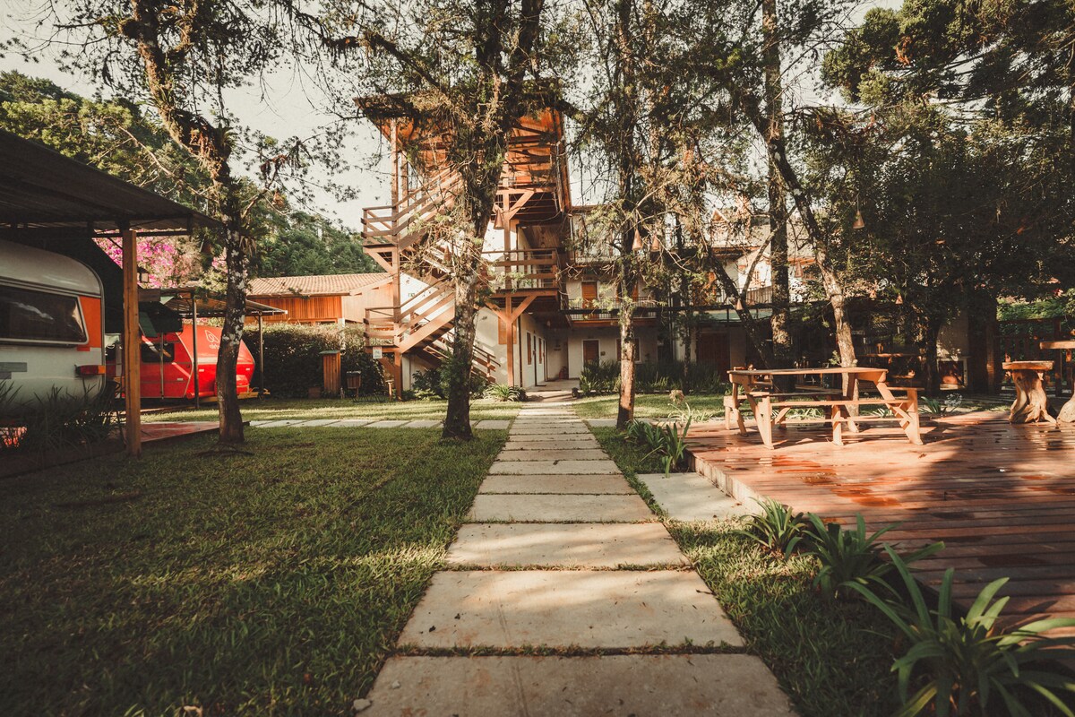 A stone pathway leads through a lush green lawn, flanked by trees and wooden seating areas. A multi-level building emerges in the background, surrounded by nature. Trailers are visible to one side, indicating a spacious and inviting outdoor environment.