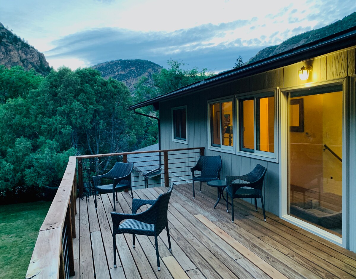 A spacious deck features several chairs arranged for relaxation, overlooking lush greenery and distant mountains. Soft evening light bathes the area, highlighting the natural surroundings while windows from the interior allow a glimpse of warm light inside.