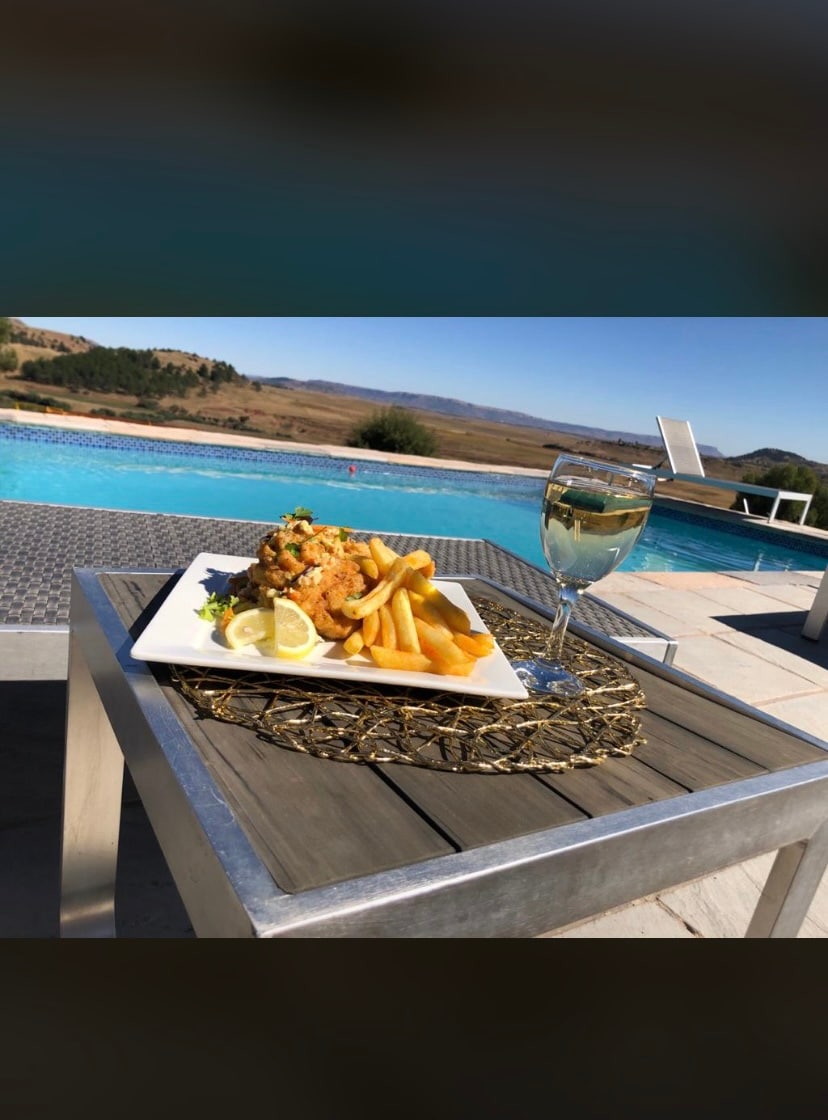 A plate of battered fish is presented alongside golden fries, garnished with lemon slices, atop a woven placemat. A glass of white wine is placed on a metallic table next to the dish, with a scenic pool and rolling hills visible in the background.