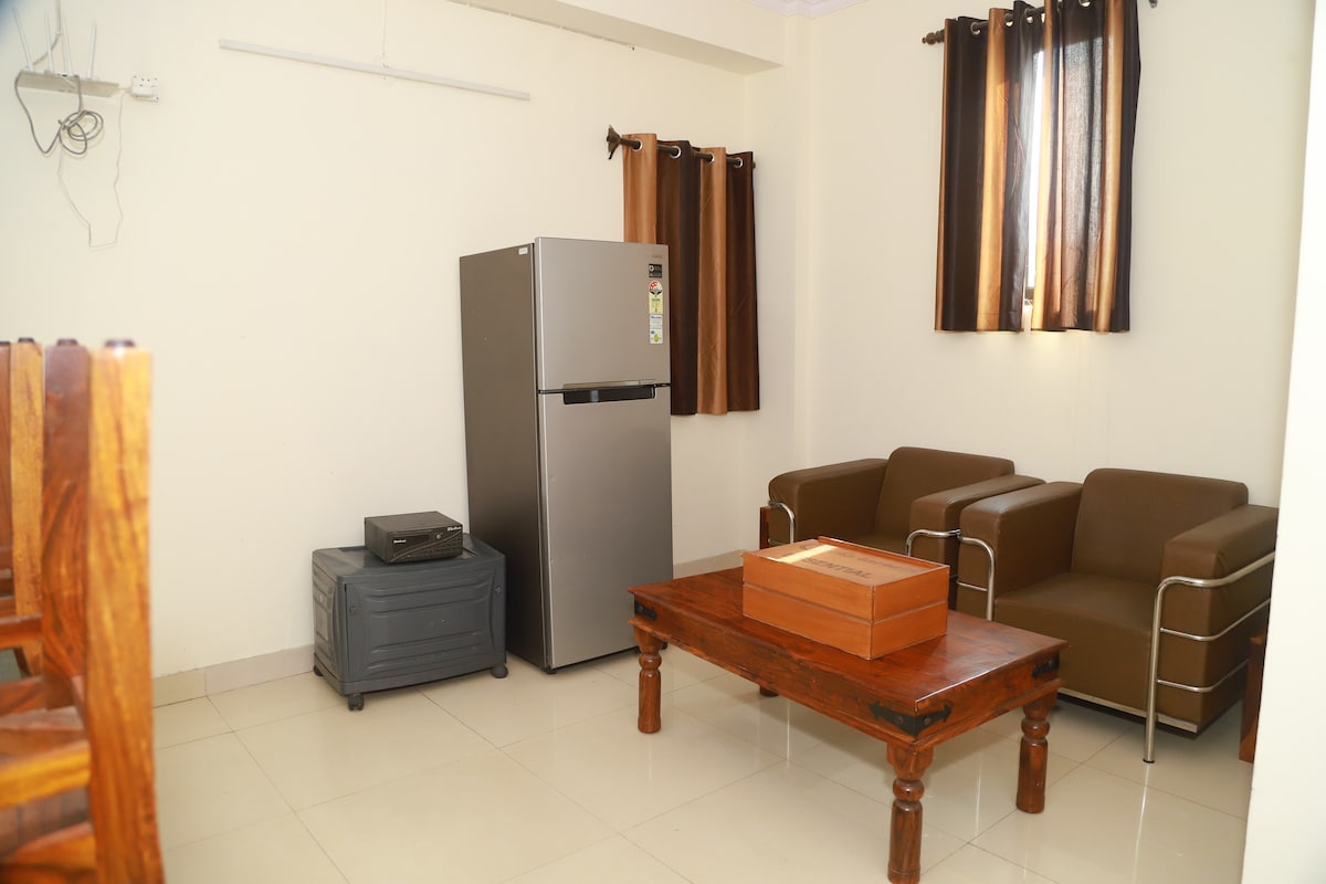 The living area features a compact seating arrangement with two brown armchairs and a wooden coffee table. A gray refrigerator stands against the wall, next to a dark blue storage box. Natural light enters through a nearby window adorned with brown curtains.