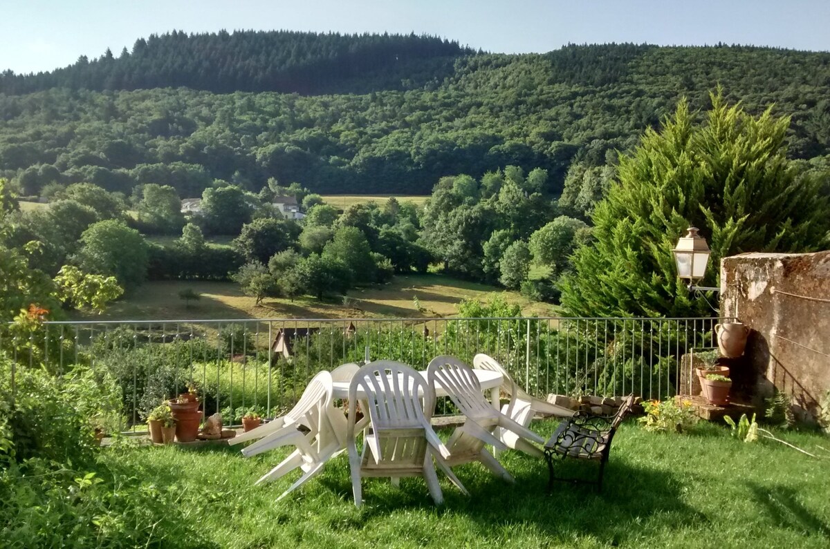 A spacious garden area features white plastic furniture arranged on green grass, with several potted plants nearby. A view of rolling hills and dense trees creates a serene backdrop, enhancing the outdoor space's inviting atmosphere.