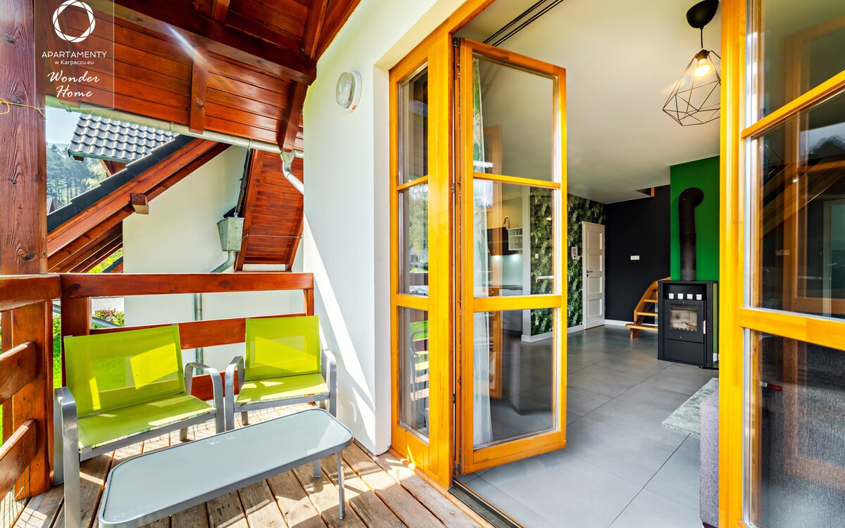 A balcony is shown, featuring two green chairs and a glass table against wooden flooring. Glass doors open into a spacious living area with a stone fireplace and a green accent wall. Natural light brightens the space, enhancing its inviting atmosphere.