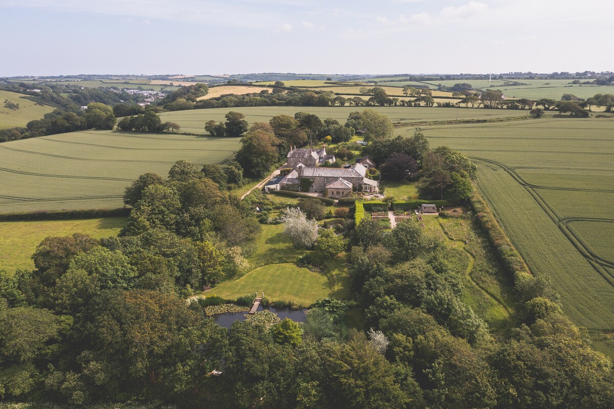 An aerial view captures the beautiful stone farm complex surrounded by lush fields and trees. The property features landscaped gardens, a serene pond, and pathways connecting various outdoor spaces, all set against a backdrop of rolling countryside in peaceful Cornish landscape.