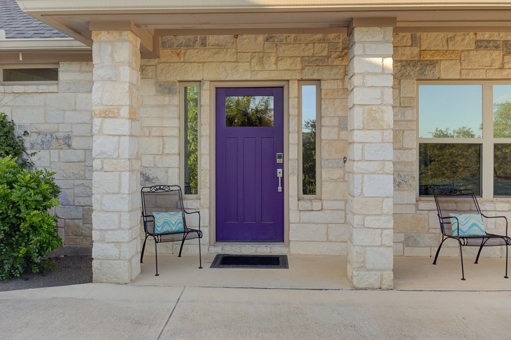 A welcoming entrance is framed by natural stone, featuring a bold purple door. Two decorative chairs are positioned on either side, providing a cozy seating area. Large windows allow natural light to brighten the entryway, blending indoor and outdoor elements harmoniously.