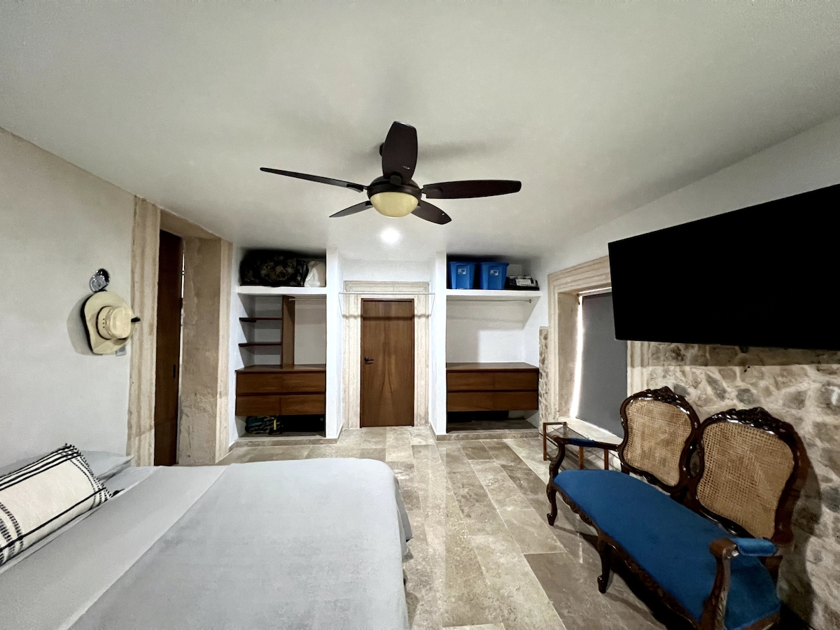 A cozy apartment is presented, featuring a spacious bed and a seating area with two wooden chairs. A ceiling fan circulates air above, while a flat-screen TV is mounted on the wall. Storage shelves and dressers are organized along one side.