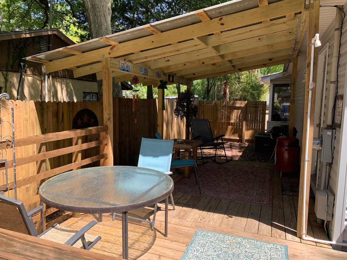 An outdoor patio area is featured, showcasing a wooden deck covered by a roof. A round glass table with four chairs is visible, complemented by a cozy seating arrangement. The space is surrounded by a wooden fence, offering privacy and a relaxed atmosphere.