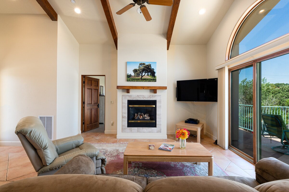 A spacious living area features vaulted ceilings with exposed wooden beams, a cozy fireplace beneath a framed artwork, and large glass doors that open to the balcony. A comfortable armchair and a wooden coffee table holding magazines and a flower vase are positioned in the room.