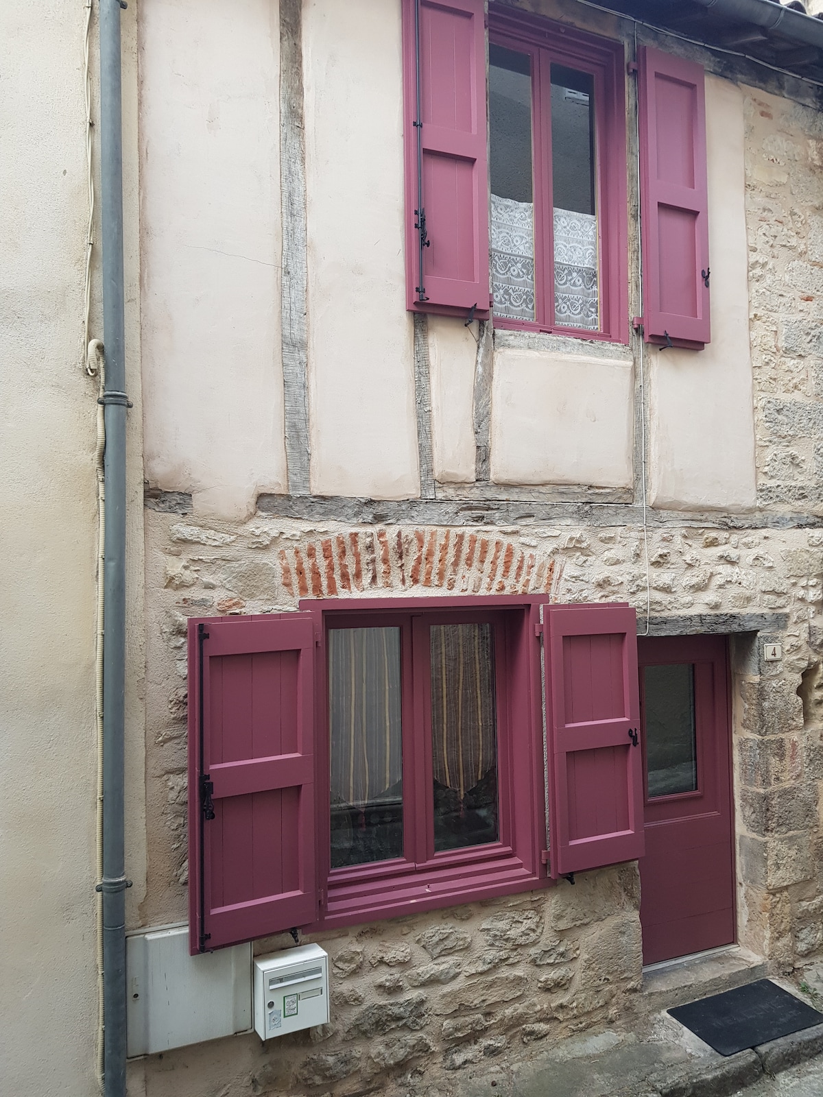 The charming exterior features a combination of stone and timber elements. Purple shutters frame the windows, creating a striking contrast against the natural stone facade. A wooden door invites entry, while the historical detailing of the architecture reflects the character of the medieval setting.