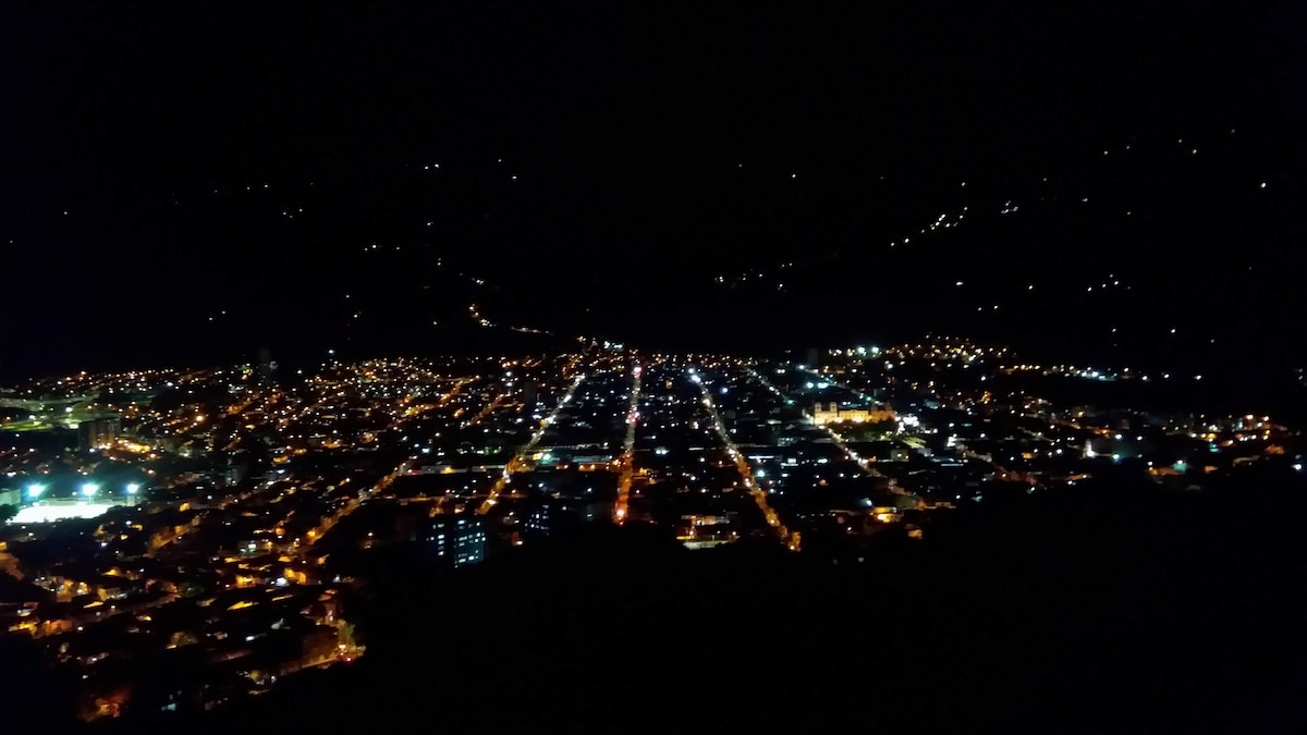 An expansive nighttime view of the city showcases a vibrant network of lights stretching across the landscape. Streets and buildings are illuminated, creating a glowing pattern against the dark backdrop of the surrounding hills.