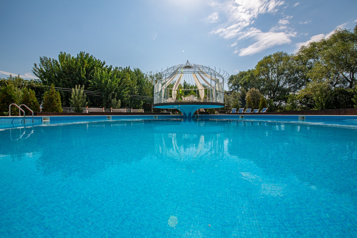 A large swimming pool features clear blue water, reflecting the sky above. White lounge chairs are positioned around the pool for guest relaxation. A gazebo is situated in the background, surrounded by greenery, enhancing the serene environment.