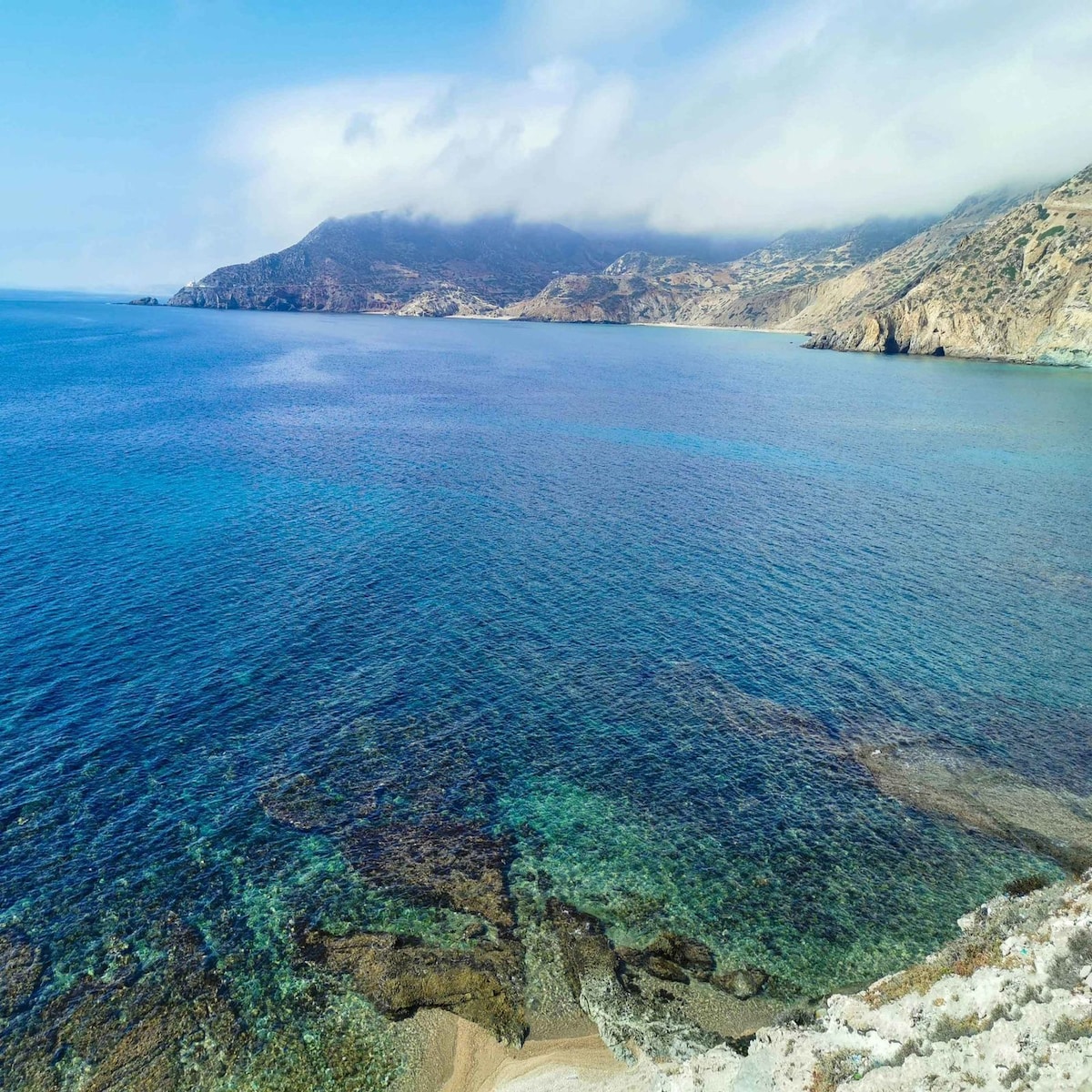 A serene coastal view features clear blue waters meeting a sandy beach, bordered by rocky cliffs and gentle waves. Soft clouds drift across a blue sky, and lush hills can be seen in the distance, creating a peaceful backdrop.