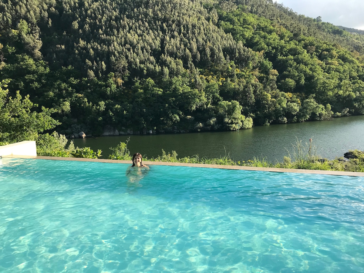 An infinity pool extends towards the river, creating a seamless view of the lush greenery of the surrounding hills. The calm water reflects the sky, enhancing the serene atmosphere of the location.
