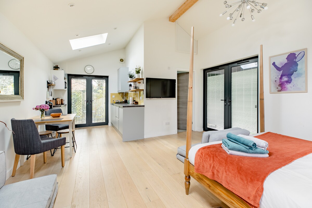 A spacious studio cabin features a four-poster bed, complemented by colorful linens. A kitchenette is visible, along with a dining area that includes wooden furniture. Large glass doors provide views of the outdoor area, while natural light illuminates the warm wood flooring.