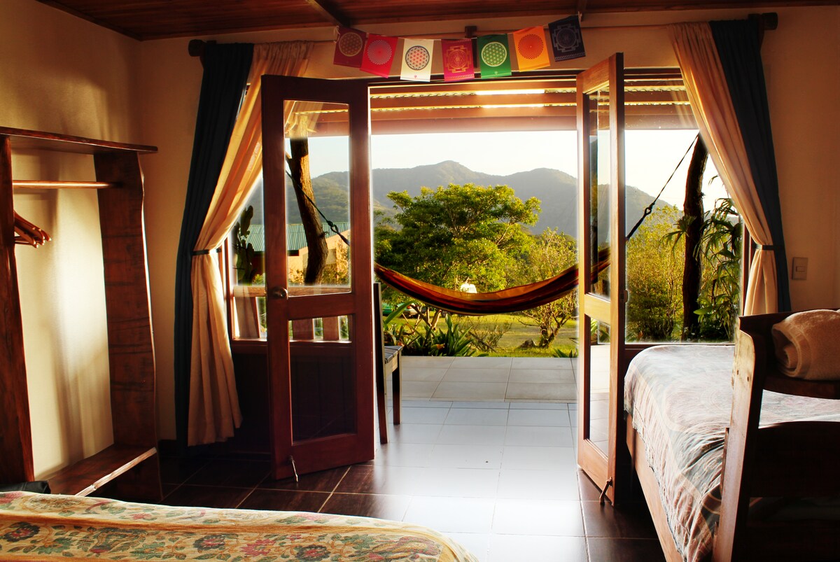 The room features large double doors that open onto a private patio with a hammock, revealing views of the mountains and surrounding greenery. Inside, a comfortable double bed and a single bed are present, complemented by wooden accents and natural light.