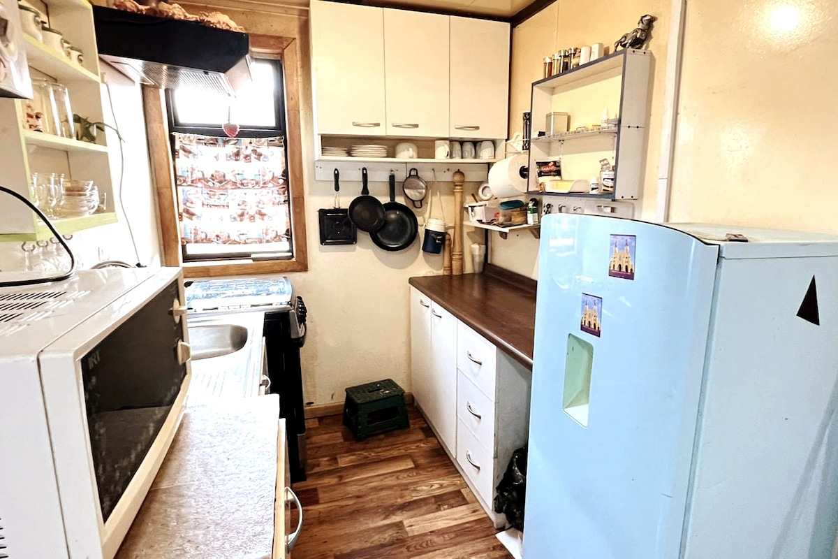 A compact kitchen is equipped with essential appliances, including a microwave and refrigerator. Cabinets and shelving provide storage for kitchenware. A wooden countertop offers workspace, with hooks mounted for hanging pots and utensils. Natural light enters through a window, enhancing the inviting atmosphere.