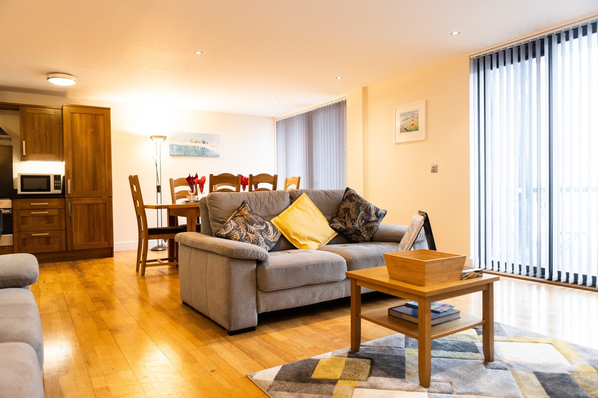 A spacious living area features a soft grey sofa adorned with colourful cushions. A wooden coffee table sits at its center. To the side, a dining table with several chairs is visible. Natural light illuminates the space through vertical blinds.