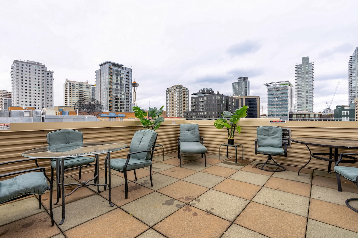 A rooftop terrace features multiple seating options, including a glass table surrounded by cushioned chairs. Potted plants add a touch of greenery against the urban backdrop of Seattle’s skyline, showcasing a mix of modern buildings and cityscape.