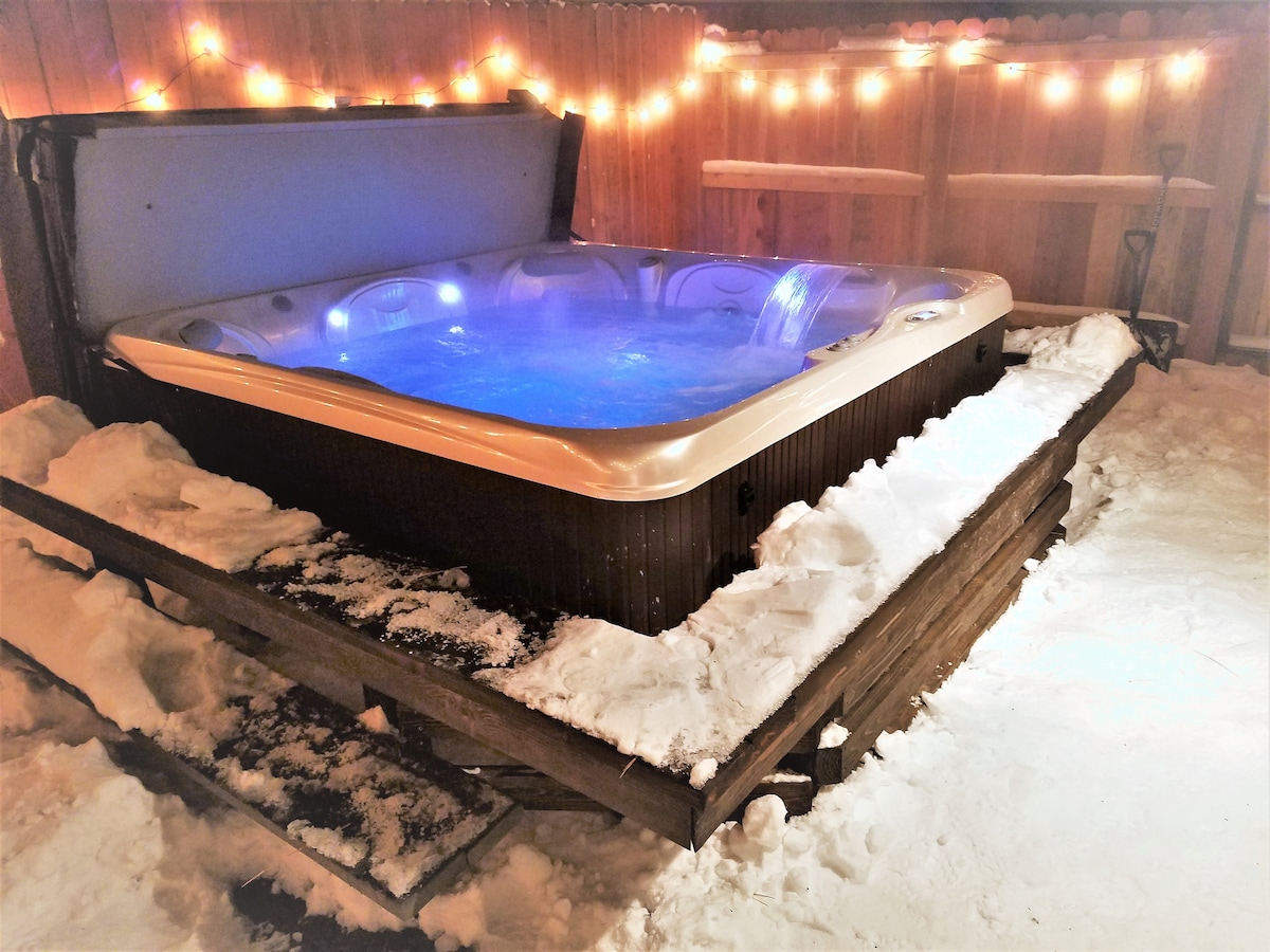A covered hot tub is surrounded by freshly fallen snow, creating a serene winter ambiance. Soft lighting from string lights above enhances the cozy setting. The tub features an inviting blue glow, suggesting a relaxing retreat after a day of outdoor activities.