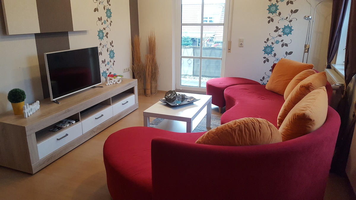 A modern living room features a curved red couch with bright orange cushions, providing a lively seating area. A light wooden TV stand holds a flat-screen television, and a white coffee table sits in the center. Large windows allow natural light to fill the space.
