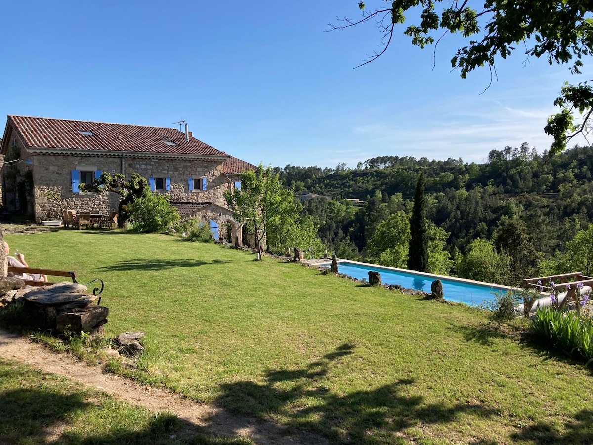 A stone house is set within a spacious grassy area, with a large swimming pool extending into the landscape. Trees and shrubs frame the property, while a clear blue sky adds to the serene atmosphere. Outdoor seating areas can be seen inviting relaxation.