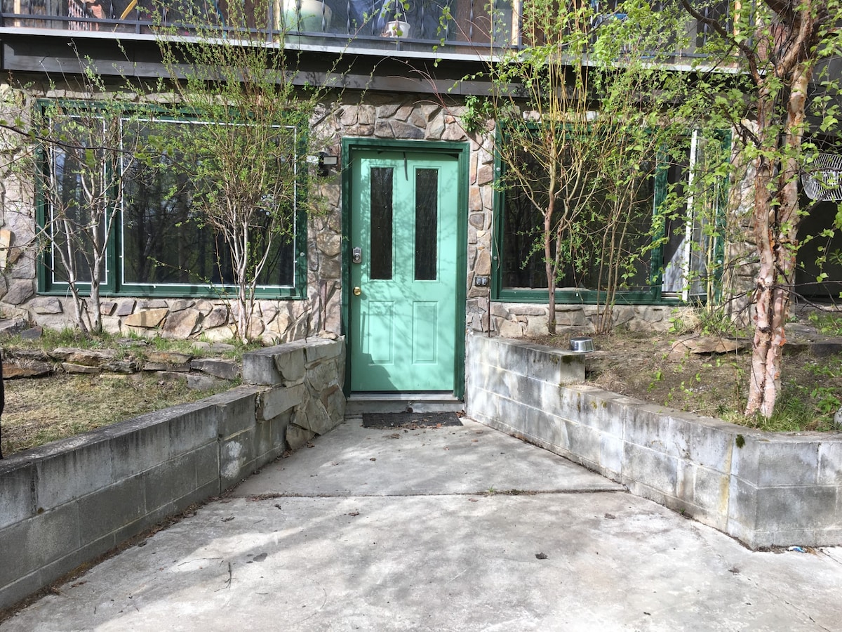 A cozy stone exterior is highlighted by a green front door, inviting guests to enter. Surrounding the entrance are landscaped areas with young trees and shrubs. A concrete pathway leads to the entrance, framed by low stone walls.