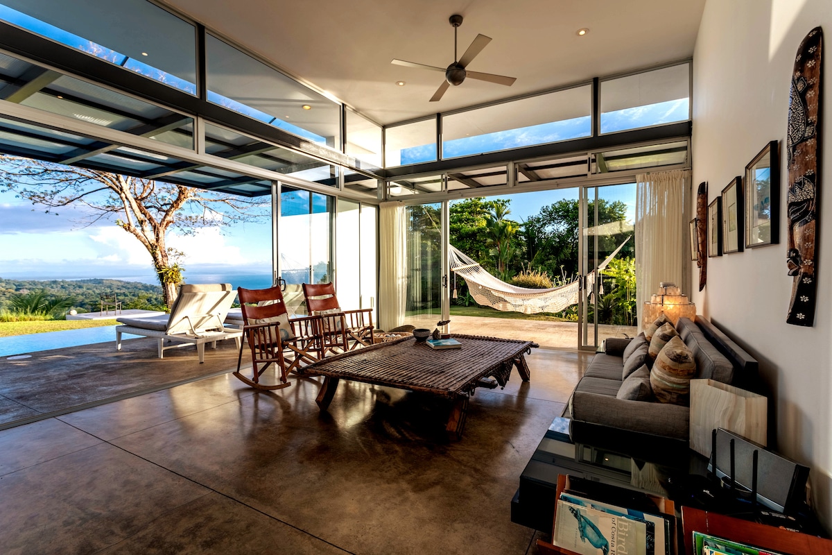 An open living area is highlighted by floor-to-ceiling glass walls, allowing natural light and views of the surrounding landscape. Comfortable seating is arranged around a low coffee table, while a hammock is visible nearby, offering a space for relaxation.