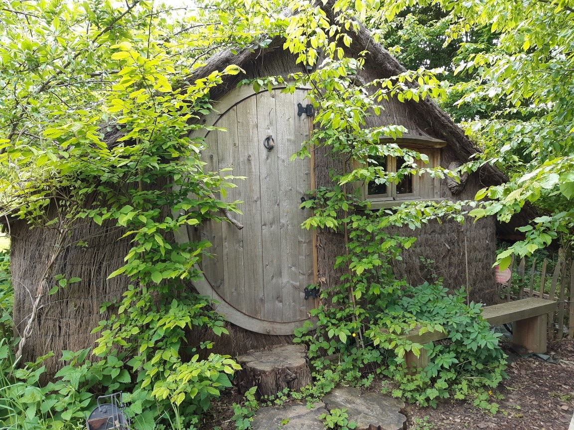 The exterior of a charming small dwelling, constructed with natural materials, is surrounded by lush greenery. A round wooden door is set into the thatched facade, framed by vibrant leaves and shrubs, while a rustic bench sits on a stone pathway in front.