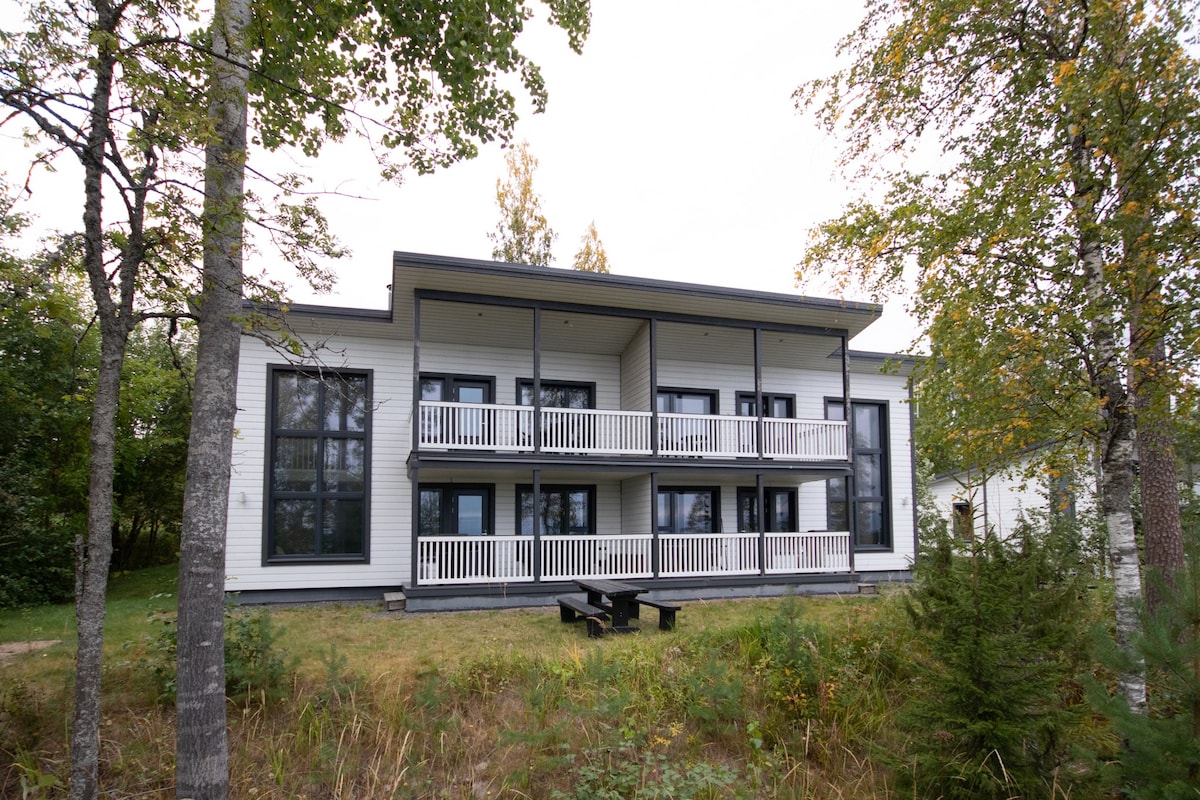 A modern two-story building is framed by trees, featuring balconies on both levels with white railings. The structure has a symmetrical layout and large windows that provide ample natural light. A grassy area with benches is visible in the foreground.