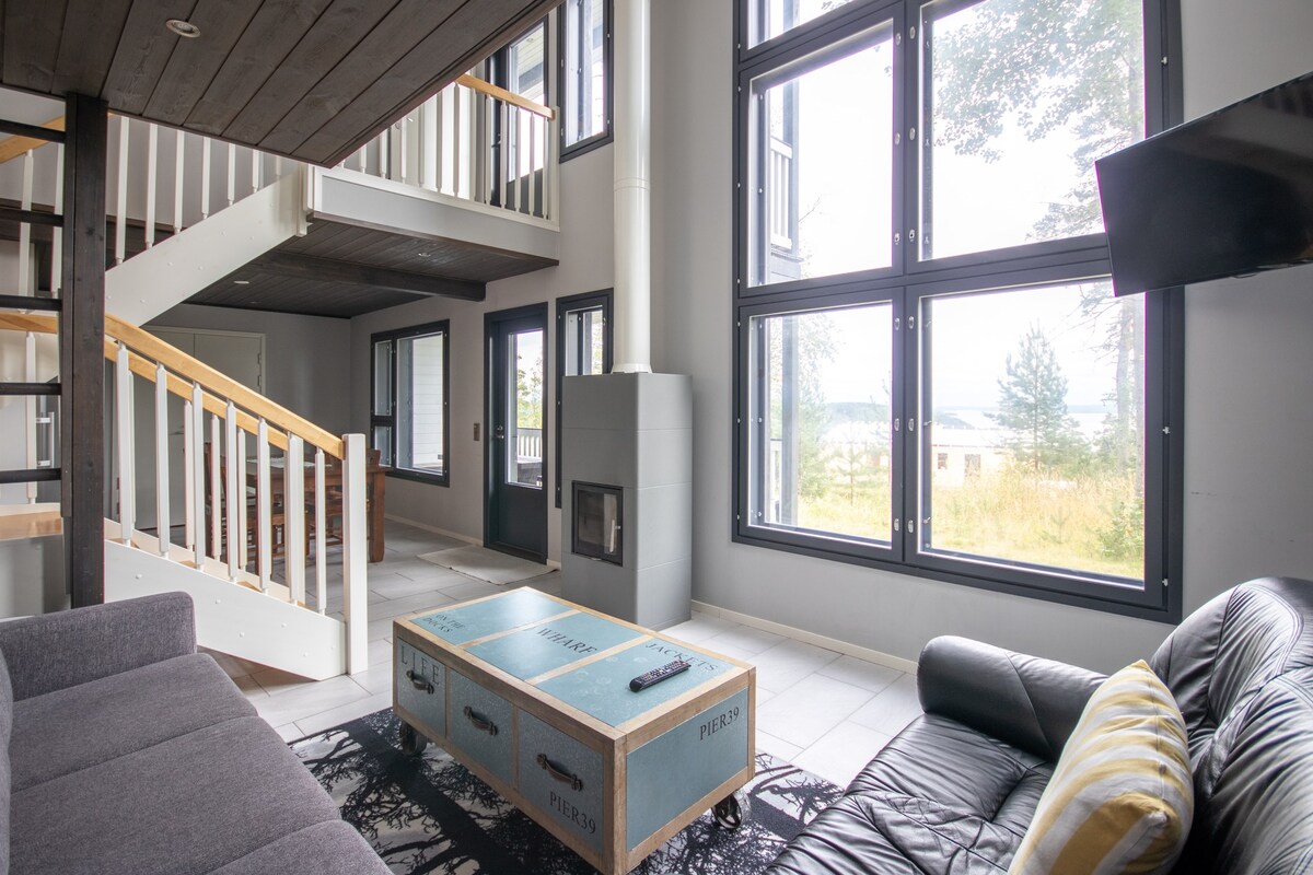 A light-filled living space features large windows that provide views of the surrounding nature. A sleek black sofa is paired with a coffee table, and a small fireplace is positioned near the seating area. A staircase leads to the second level, enhancing the open design.
