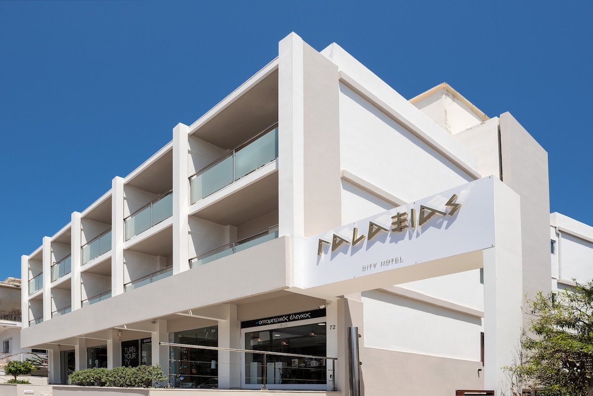 The hotel exterior features a modern white façade with multiple balconies. Large glass windows allow natural light to fill the interior. The building is complemented by a clear blue sky, enhancing the contemporary architectural design of this welcoming accommodation.