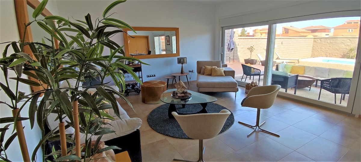 A spacious living area features a large glass door that opens to the garden and swimming pool. Comfortable seating is arranged around a round coffee table on a textured rug. Natural light fills the room, complemented by indoor plants and modern decor.