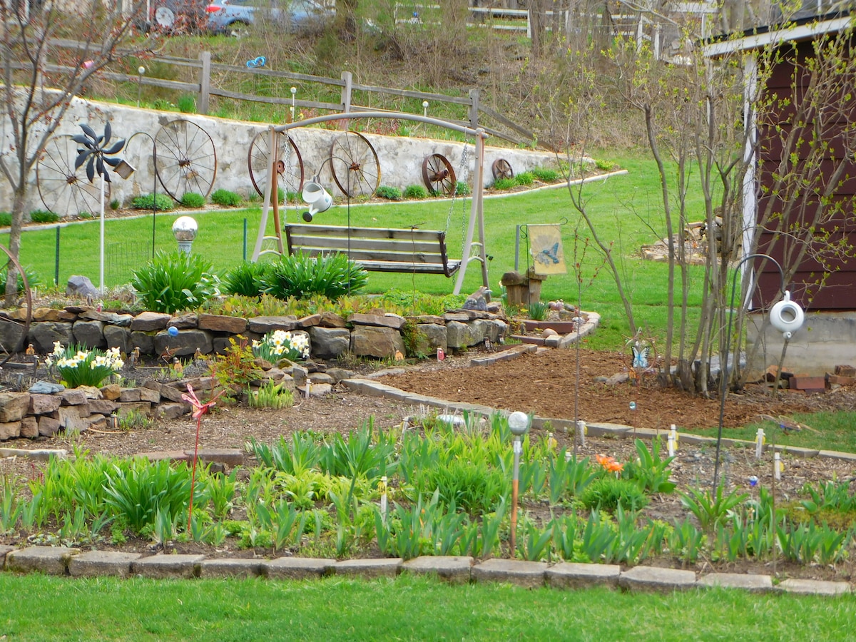 A landscaped garden features a variety of perennial plants and flowers, with a stone border outlining the flower beds. A swing is placed among the greenery, and decorative items are scattered throughout. Rolling hills and a wooden fence create a tranquil backdrop.