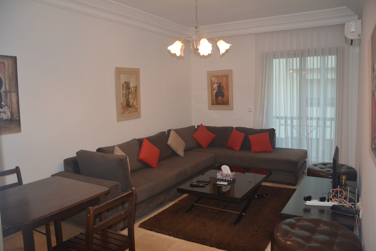 A spacious living area is seen, featuring a large L-shaped sofa adorned with red accents. A glass coffee table sits atop a soft area rug, providing a cozy focal point. Natural light flows through the window, complemented by sheer curtains. Wall art adds visual interest.