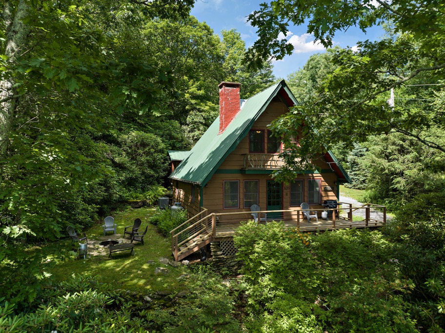 A charming cabin is nestled among lush greenery, showcasing a sloped roof with green accents and a welcoming porch. The outdoor seating area is surrounded by trees, creating a serene atmosphere. The building's wooden façade complements the natural setting.