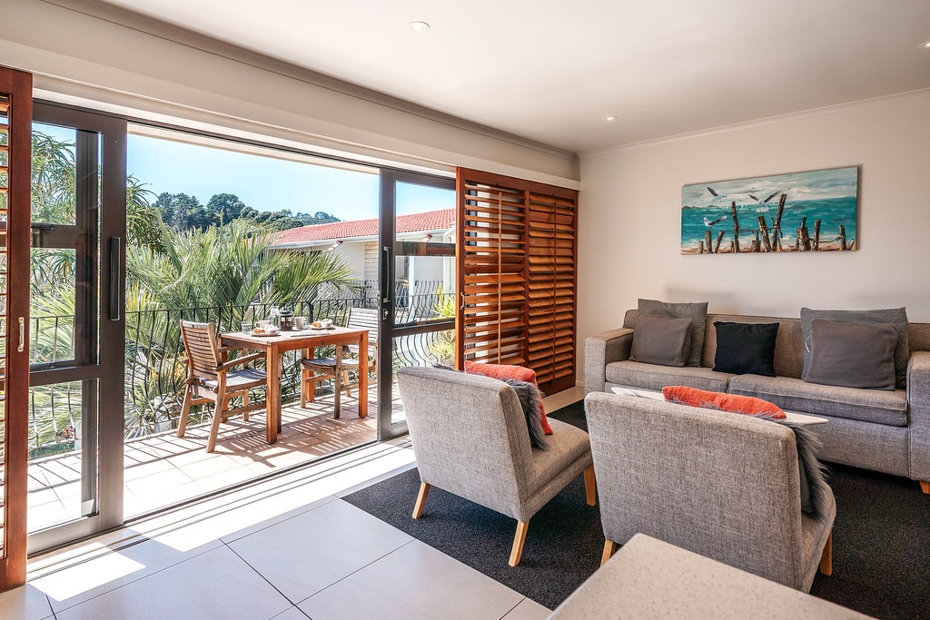 A light-filled living area features large sliding doors leading to an outdoor balcony. A wooden dining table accommodates two, while a comfortable sofa with cushions invites relaxation. Various plants are visible outside, enhancing the view of the landscaped courtyard.